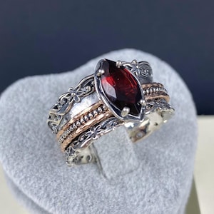 May include: A silver ring featuring a red gemstone and intricate floral designs. The ring has a rose gold band and is set with a marquise-shaped gemstone.
