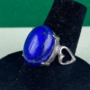 Op de afbeelding: Een zilveren ring met een grote ovale lapis lazuli-edelsteen. De ring heeft een hartvormig ontwerp op de band.
