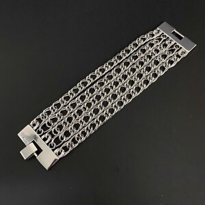 May include: Silver bracelet with five parallel chain strands. The bracelet has a rectangular clasp at each end. The chains are made of interlocking oval links. The bracelet is displayed on a black surface.