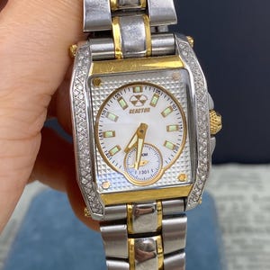 May include: A rectangular Reactor watch with a white face, gold accents, and a silver band. The watch face features a smaller dial and the word "REACTOR". The bezel is encrusted with small, clear stones.