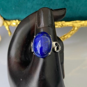 Op de afbeelding: Een zilveren ring met een grote ovale blauwe lapis lazuli edelsteen. De ring is geplaatst op een zwarte achtergrond.