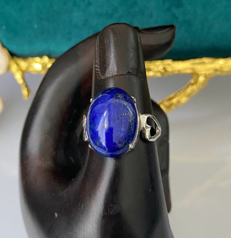 Op de afbeelding: Een zilveren ring met een grote ovale blauwe lapis lazuli edelsteen. De ring is geplaatst op een zwarte achtergrond.