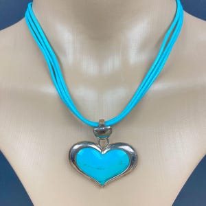 Puede incluir: Un collar con colgante de corazón turquesa con borde plateado y un cordón azul brillante. El colgante en forma de corazón está relleno de una vibrante piedra turquesa. El collar se exhibe en un maniquí.