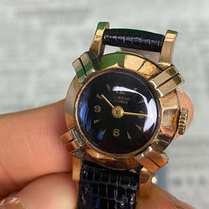 May include: Vintage Geneva watch with a black face, gold-tone case, and black leather band. The watch face displays the numbers 6 and 9. The watch has a rectangular black leather band. The watch is held by a person.