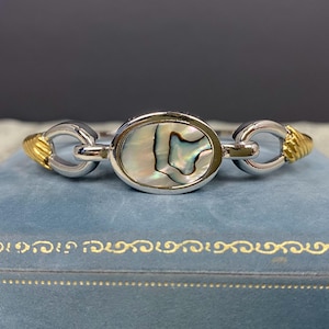 May include: Silver and gold-tone bracelet featuring an oval abalone shell centerpiece. The bracelet has a silver chain link design with gold-tone accents on each end. The abalone shell has iridescent colors of green, blue, and white.
