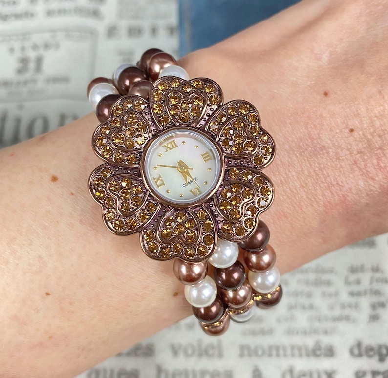 May include: A flower-shaped quartz watch with a bronze-colored bezel and gold-colored crystals. The watch face is white with gold hands and Roman numerals. The band is made of brown and white beads.