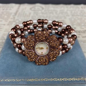 May include: A flower-shaped watch with a pearl and bead bracelet. The watch face is white with gold-tone hands and Roman numerals. The flower-shaped watch case is adorned with small, sparkling crystals. The bracelet features white, brown, and bronze-colored beads.