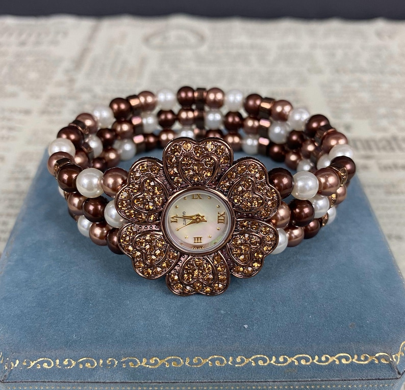 May include: A flower-shaped watch with a pearl and bead bracelet. The watch face is white with gold-tone hands and Roman numerals. The flower-shaped watch case is adorned with small, sparkling crystals. The bracelet features white, brown, and bronze-colored beads.