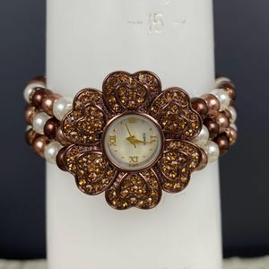 May include: A floral-shaped quartz watch with a cream-colored face and gold hands. The watch is surrounded by a flower design with brown and gold rhinestones. The band is made of white and brown beads.