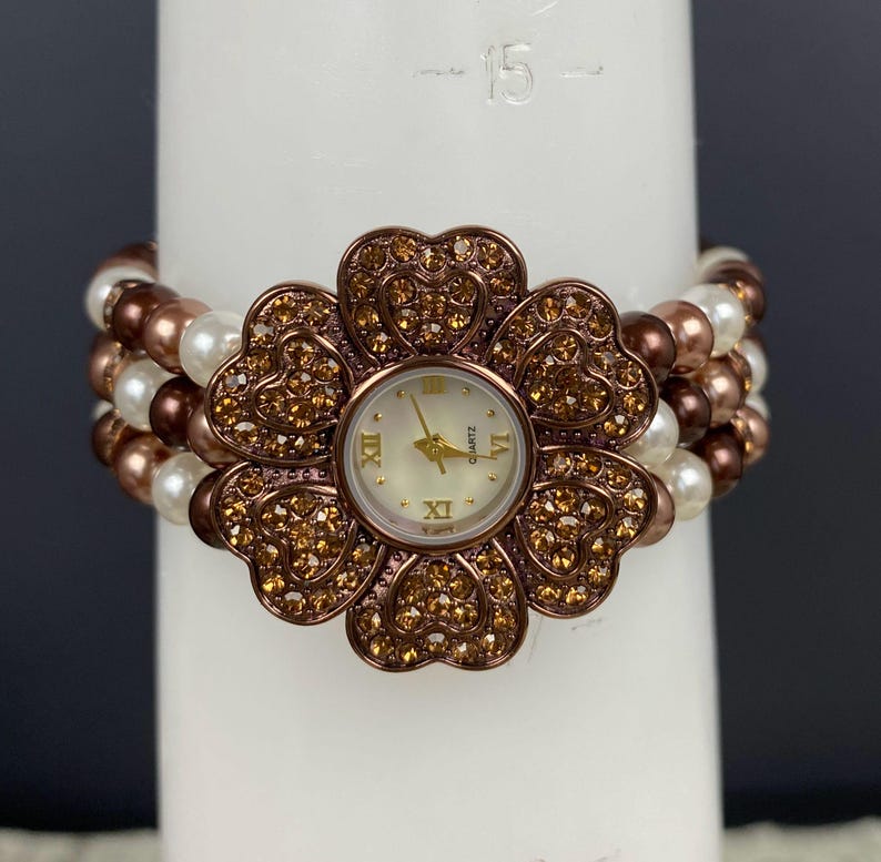 May include: A floral-shaped quartz watch with a cream-colored face and gold hands. The watch is surrounded by a flower design with brown and gold rhinestones. The band is made of white and brown beads.