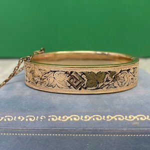 May include: A gold-coloured bangle bracelet featuring detailed floral and geometric patterns in black and green. The bracelet has a hinged clasp and a safety chain. The bracelet is on a blue surface.