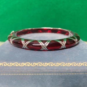 May include: A hinged bangle bracelet featuring a series of deep red, translucent, curved segments separated by polished silver-toned metal X-shaped accents. The bracelet has a smooth, rounded profile and a secure clasp mechanism visible on the side.