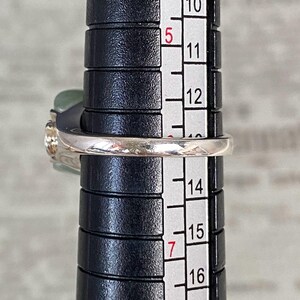 May include: A silver ring with a light green stone and a clear stone is being measured on a black ring sizer. The ring is between sizes 13 and 14 on the sizer. The ring has a delicate design.
