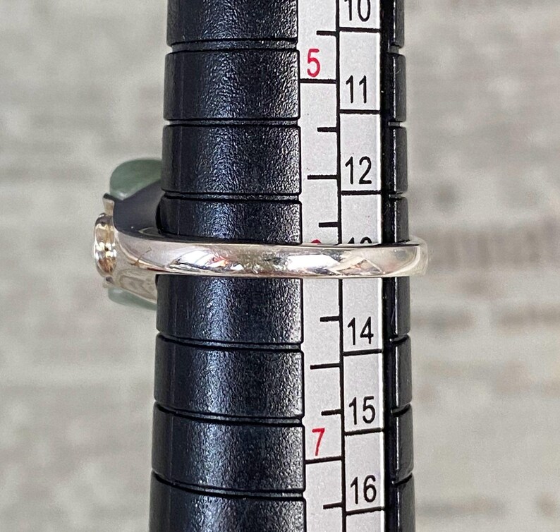 May include: A silver ring with a light green stone and a clear stone is being measured on a black ring sizer. The ring is between sizes 13 and 14 on the sizer. The ring has a delicate design.