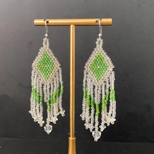 May include: A pair of long, beaded earrings with a green and white diamond pattern. The earrings are hanging from a gold stand.