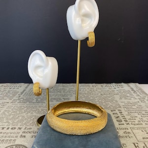 Peut inclure: Ensemble de bijoux dorés comprenant un bracelet jonc texturé et des créoles assorties. Les boucles d'oreilles sont présentées sur des présentoirs blancs en forme d'oreille. Le bracelet repose sur un coffret en velours bleu, mettant en valeur le design et la couleur des bijoux.