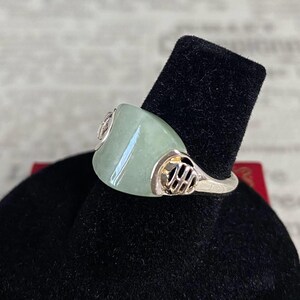 May include: A silver ring with a large, light green jade stone. The ring has decorative silver accents on the sides of the jade. The ring is displayed on a black velvet surface.