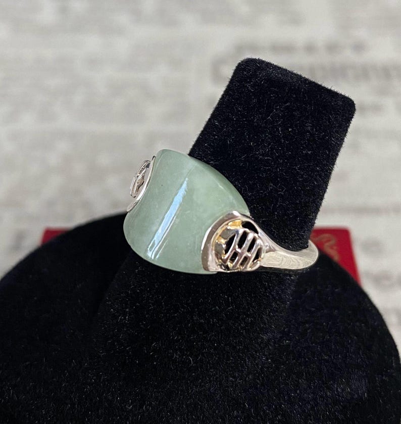 May include: A silver ring with a large, light green jade stone. The ring has decorative silver accents on the sides of the jade. The ring is displayed on a black velvet surface.
