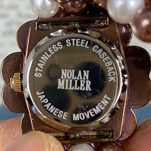 May include: Close-up of a Nolan Miller watch. The watch has a copper-colored, flower-shaped case and a stainless steel caseback. The caseback reads "Stainless Steel Caseback", "Nolan Miller", and "Japanese Movement". The watch is adorned with pearl beads.