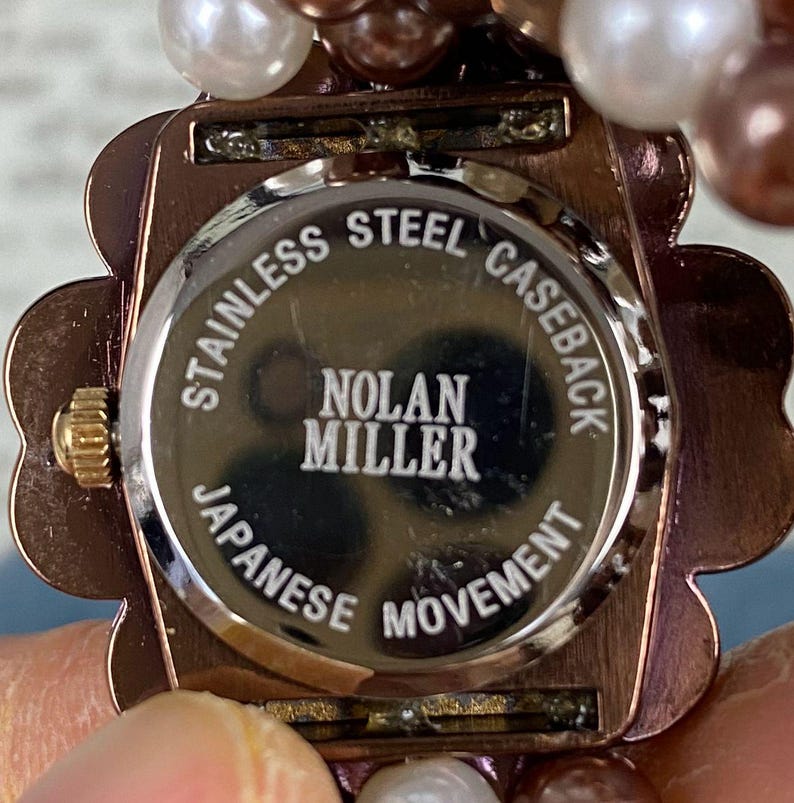 May include: Close-up of a Nolan Miller watch. The watch has a copper-colored, flower-shaped case and a stainless steel caseback. The caseback reads "Stainless Steel Caseback", "Nolan Miller", and "Japanese Movement". The watch is adorned with pearl beads.