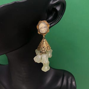 Może przedstawiać: Złoty kolczyk w stylu vintage z perłowym, owalnym wierzchem i ażurowym wzorem stożka. Poniżej stożka zwisa kilka jasnozielonych, nieregularnych koralików. Kolczyk jest prezentowany na czarnej głowie manekina na zielonym tle.