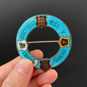 Könnte beinhalten: Eine runde Brosche aus blauem Emaille mit vier Wappen und dem Text "SOIT QUI MALY PENSE" am Rand.