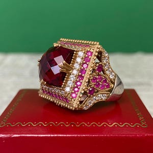 May include: A large, ornate ring with a faceted, deep red gemstone as the centerpiece. The ring features gold filigree and is encrusted with smaller red and white gemstones. The band is silver. The ring is displayed on a red surface.