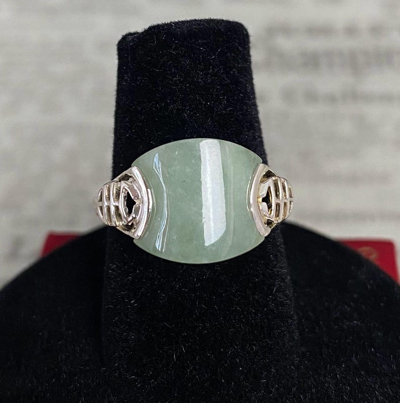 May include: A light green jade ring with a silver band. The jade is oval-shaped and set in a silver band with decorative filigree on each side. The ring is displayed on a black velvet ring holder.