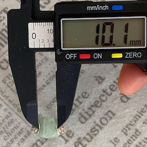 May include: A light green jade bead, approximately 0.4 inches in diameter, is measured with a digital caliper. The caliper displays a measurement of 10.1 mm. The bead is held between the caliper jaws, showing its size.