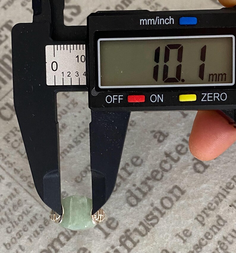 May include: A light green jade bead, approximately 0.4 inches in diameter, is measured with a digital caliper. The caliper displays a measurement of 10.1 mm. The bead is held between the caliper jaws, showing its size.
