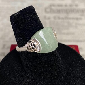May include: A silver ring with a light green, rectangular jade stone. The ring has intricate silver filigree designs on the sides and a polished finish. The jade stone is smooth and has a slight sheen, set on a black velvet display.