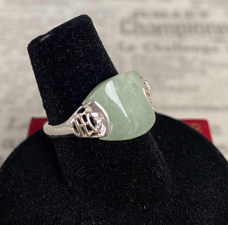 May include: A silver ring with a light green, rectangular jade stone. The ring has intricate silver filigree designs on the sides and a polished finish. The jade stone is smooth and has a slight sheen, set on a black velvet display.
