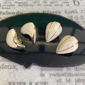 Könnte beinhalten: Vier Vintage-Ohrringe. Zwei sind halbmondförmig mit cremefarbenem Emaille und goldfarbenen Zierleisten. Die anderen beiden sind tropfenförmig mit cremefarbenem Emaille und goldfarbenen Zierleisten. Die Ohrringe werden auf einer dunkelgrünen und schwarzen Oberfläche präsentiert.