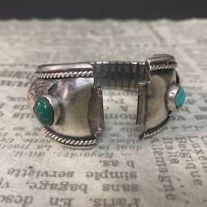 May include: A silver-toned cuff bracelet featuring two round turquoise stones set on either side of the opening. The metal surface displays a polished finish with twisted rope detailing along the edges and a leaf engraving near one stone.