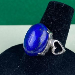 Op de afbeelding: Een zilveren ring met een grote ovale lapis lazuli-edelsteen en een hartvormig ontwerp op de band.