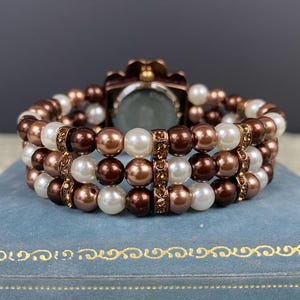 May include: A multi-strand bracelet featuring brown, white, and bronze-colored faux pearls. The bracelet has a watch face with a bronze-colored floral design. The bracelet is resting on a blue surface.