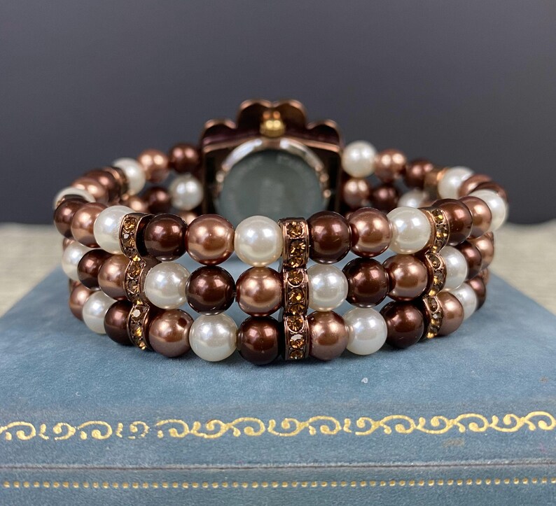 May include: A multi-strand bracelet featuring brown, white, and bronze-colored faux pearls. The bracelet has a watch face with a bronze-colored floral design. The bracelet is resting on a blue surface.