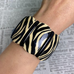May include: A wide, bold bracelet with a zebra stripe pattern. The bracelet features alternating black and cream-colored curved rectangular links. The bracelet is worn on a wrist, showcasing its design and style.