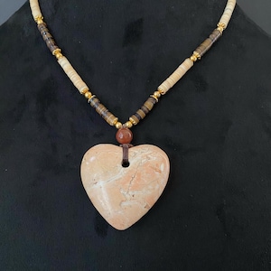 May include: A heart-shaped pendant necklace with a light pink and white marbled stone. The necklace is made with brown and beige beads and gold accents.