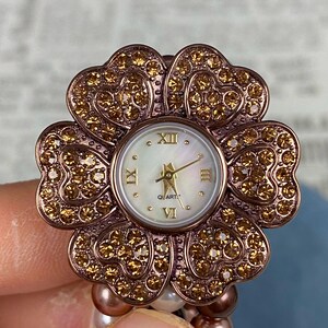 May include: A flower-shaped watch with a rose gold finish and sparkling crystals. The watch face is white with gold hands and Roman numerals. The watch is attached to a beaded bracelet.