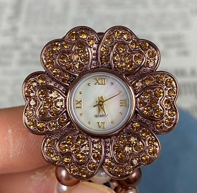 May include: A flower-shaped watch with a rose gold finish and sparkling crystals. The watch face is white with gold hands and Roman numerals. The watch is attached to a beaded bracelet.