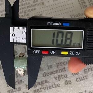May include: A digital caliper measuring a jade ring. The ring has a light green oval stone and a gold-colored band. The digital display shows a measurement of 10.8 mm.