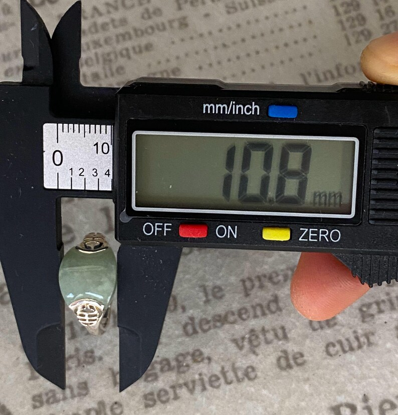 May include: A digital caliper measuring a jade ring. The ring has a light green oval stone and a gold-colored band. The digital display shows a measurement of 10.8 mm.