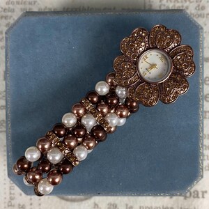 May include: A flower-shaped watch with a white face and gold hands. The watch is adorned with brown rhinestones and sits atop a band of brown, white, and gold beads. The watch is a stylish accessory.