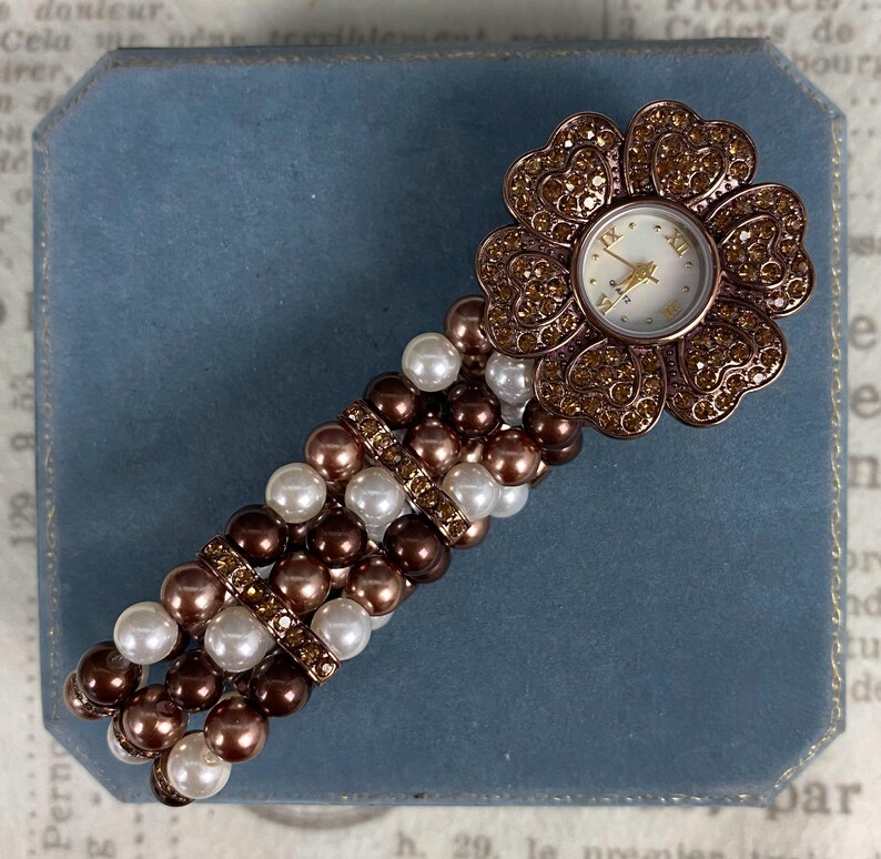 May include: A flower-shaped watch with a white face and gold hands. The watch is adorned with brown rhinestones and sits atop a band of brown, white, and gold beads. The watch is a stylish accessory.