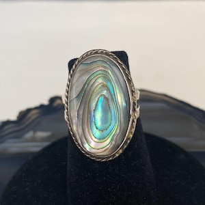 May include: Oval-shaped abalone shell ring with a silver-tone rope-like border. The iridescent shell displays concentric patterns of blue, green, and white. The ring is set on a black display stand.