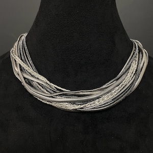 May include: A multi-strand necklace featuring a collection of intertwined metallic chains in varying shades of silver and dark gunmetal gray. The design consists of smooth snake chains and textured braided strands layered together in a circular form.