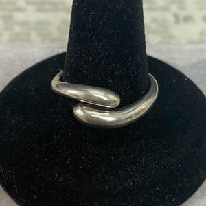 May include: A polished silver-tone bypass ring featuring two smooth, rounded, tapered ends that overlap slightly. The metal has a reflective, metallic finish with a curved, fluid design that wraps around the finger, showcasing a simple, continuous band.