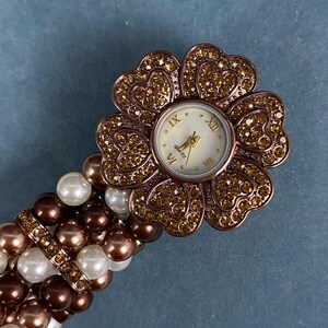 May include: A flower-shaped quartz watch with a white face and gold hands. The watch is surrounded by sparkling crystals and is attached to a bracelet made of white, brown, and pearl-colored beads. The watch face displays Roman numerals.