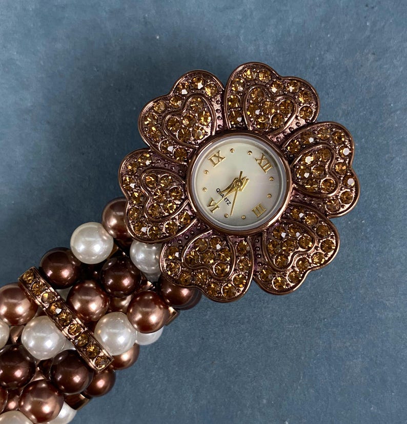 May include: A flower-shaped quartz watch with a white face and gold hands. The watch is surrounded by sparkling crystals and is attached to a bracelet made of white, brown, and pearl-colored beads. The watch face displays Roman numerals.